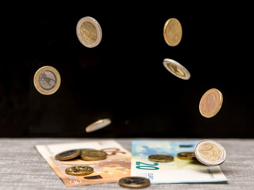 In this image, various Euro coins, including one- and two-euro pieces with metallic silver and gold finishes, are shown mid-air above a flat surface, with some coins already resting on cash, such as a ten-euro banknote and a five-euro banknote, partially visible at the bottom. The coins display different textures, with some having ridged edges and others smooth, and their surfaces reflect light against a dark background, creating a sense of motion as the coins appear to be falling or being tossed. The banknotes have distinctive colours and patterns typical of Euro currency, with visible numbers and symbols. The scene is set on a grey fabric surface, likely a table or countertop. This visual representation aligns with themes of financial transaction or the handling of money, which could be related to private waste disposal or alternative investment in waste clearance services, such as those provided by Waste Clearance Merton, to support efficient rubbish removal and avoid illegal fly-tipping.