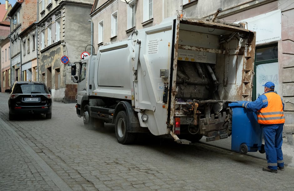 A row of five large wheelie bins lined up against a plain, light-colored wall outdoors. The bins are made of dark grey plastic with textured surfaces and sturdy, smooth yellow lids. Each bin has a small white oval label with black text, possibly indicating identification or collection details, affixed to the front. The bins are positioned on a concrete surface, with the minimal shadow indicating daylight. The background wall is smooth and unadorned, emphasizing the uniform appearance of the bins, which are arranged neatly side by side. The scene reflects a typical communal or private waste collection setup, possibly associated with a rubbish removal service such as Waste Clearance Merton, highlighting an organized approach to domestic or commercial waste storage and collection.