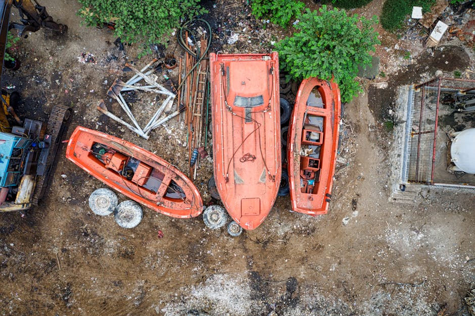 An aerial view of a construction or industrial site showing three small red boats and a large orange utility vehicle parked on uneven dirt ground. The boats, positioned side by side, feature visible interiors with some equipment and seating, and are weathered with paint peeling in places. The larger orange vehicle, which appears to be a type of loader or utility truck, is centrally located and shows signs of wear with dirt and minor rust visible on its surface. Surrounding the boats and vehicle, there are scattered metal and plastic components, including pipes, panels, and containers, alongside loose gravel and dirt. To the left side of the image, a small piece of construction machinery with tracks can be seen, and behind the equipment, there is a patch of green foliage, including trees and shrubs, providing some natural contrast to the industrial scene. On the right, part of a brick wall and a small fenced area are visible, suggesting the site is enclosed. This scene illustrates a storage or disposal area for older or unused equipment, aligning with the context of private waste handling and rubbish removal services provided by Waste Clearance Merton.