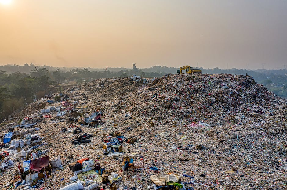 A large open landfill site filled with a vast mound of mixed waste materials, including plastics, textiles, paper, and tires, which are scattered across the uneven terrain. The waste is piled high, with some larger items like bundles of trash and discarded furniture nestled within the heap, and smaller debris such as plastic bottles and packaging spread throughout. The surface appears rough and irregular, with varying textures from soft fabrics to hard plastics and rubber. In the background, distant trees and faint structures are visible under a hazy sky, possibly due to atmospheric pollution or dust from the landfill. To the right of the mound, a yellow excavator is positioned atop the waste, indicative of active loading or management activities. The scene captures an outdoor environment typical of waste disposal or clearance operations, highlighting the scale of waste accumulated at such sites without any human presence in the immediate foreground. Waste Clearance Merton may handle local rubbish removal and associated waste management, demonstrating the importance of proper disposal methods for large-scale refuse accumulation.