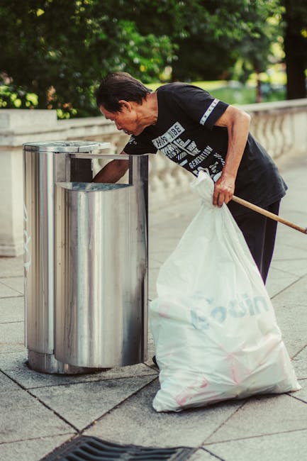 A middle-aged man with short dark hair wearing a black T-shirt with white text is seen disposing of a white rubbish bag into a modern, stainless steel litter bin situated on a paved sidewalk alongside a low stone wall. The stainless steel bin has a smooth, reflective surface and an open top, designed for easy waste disposal. The scene is set outdoors in a well-maintained area with lush green trees and foliage visible in the background, suggesting a park or public space. The man is holding a long stick or litter picker in his left hand, which is used to help deposit the bag into the bin, emphasizing a focus on cleanliness and waste management in public areas. The environment appears to be well-lit by natural daylight, enhancing the clarity of the scene. This image underscores the importance of individual responsibility in waste disposal, aligning with services offered by companies like Waste Clearance Merton for private or commercial rubbish removal outside of municipal collection services.