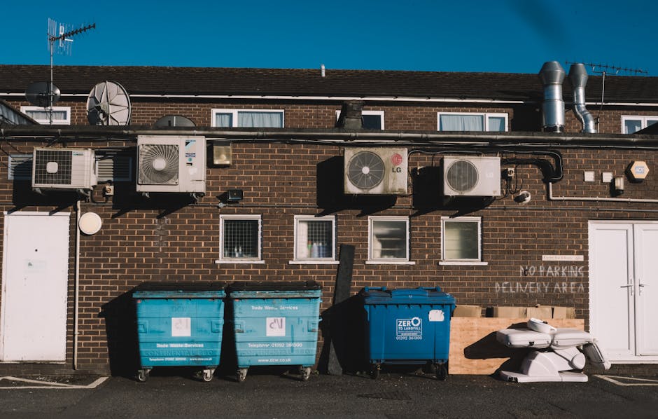 This image shows the rear exterior wall of a brick building with several mounted air conditioning units and ventilation pipes along the upper section. In the foreground, there are three large waste receptacles on a paved surface; two are blue recycling bins with white labels, and one is a darker blue bin with a white logo, all placed side by side against the wall. A white, wheeled, possibly plastic or metal object, is positioned to the right of the bins. The background features a clear blue sky, and nearby windows and a door are visible on the building's brick facade. Environmental details include the pavement and a small section of signage, indicating designated waste collection or disposal area, with subtle references to private waste management services such as those provided by Waste Clearance Merton. The scene is well-lit by natural daylight, emphasizing the textures of the brick wall, metal equipment, and wheeled bins, all contributing to a typical urban waste management setting suitable for rubbish collection and on-site clearance services.