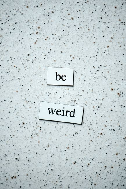 The image shows a speckled, light-colored surface, possibly a terrazzo or concrete floor, with two white rectangular tiles placed on it. The tiles have black lowercase text, with the top tile displaying the word 'be' and the lower tile showing the word 'weird.' The tiles are positioned slightly diagonally from each other, with the word 'weird' appearing below and to the right of 'be.' The environment suggests an indoor setting, and the focus on the tiles creates a contrast against the textured background, evoking a subtle message that could be linked to themes of perception or attitude, relevant to privately managed waste disposal or alternative rubbish handling options provided by Waste Clearance Merton.