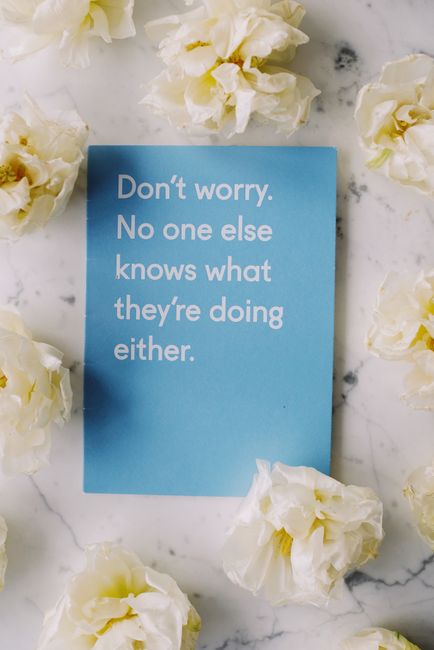 A light blue card with the message 'Don't worry. No one else knows what they're doing either.' printed in white text, positioned on a smooth marble surface with subtle grey veining. The card is surrounded by scattered cream-colored flowers with ruffled petals, some resting partially on the marble and others overlapping the edges of the card. The overall scene has a soft, natural lighting that highlights the textures of the flowers and the reflective quality of the marble, creating a calm and subtly humorous visual. This setting suggests a casual, friendly atmosphere that aligns with themes of reassurance and informal communication, potentially tying into the concept of alternative or private approaches to waste management or service options, as offered by companies like Waste Clearance Merton.