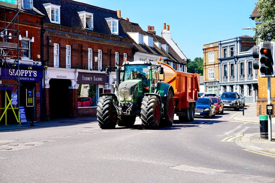 A large green tractor with black tires and a white roof is positioned at the intersection of a busy urban street, with an orange waste collection container attached to its rear. The vehicle is in motion or stopped briefly, surrounded by parked cars along the side of the road. To the left, a storefront with a sign reading 'Sloppy’s Sports Bar' and a purple awning is visible, alongside a salon with the sign 'Tierney Salons.' The buildings are primarily brick structures, featuring multiple windows, some with white frames, and a mix of residential and commercial use. The scene takes place during bright daylight, under a clear blue sky, with trees partially visible in the background. The overall setting reflects an active town or city centre where municipal or private waste collection services might operate, supporting the need for efficient rubbish removal solutions, such as those offered by Waste Clearance Merton, within the context of managing household or commercial waste.
