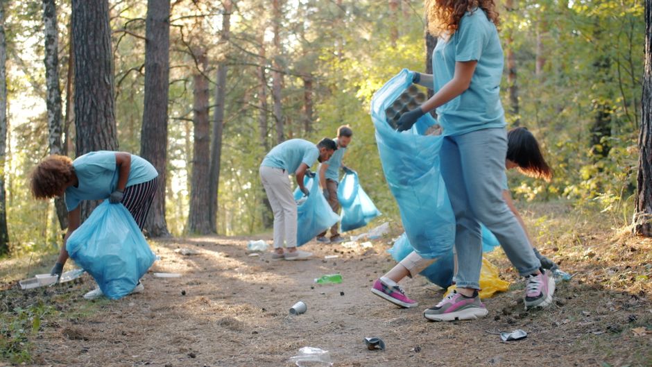 A group of volunteers wearing light blue t-shirts are participating in a litter pick-up activity in a wooded area during daylight. They are moving along a dirt path, each holding large blue trash bags filled with collected rubbish, including plastic bottles and cans. One person near the foreground is bending down to pick up litter, while others further back are actively collecting debris and placing it into their bags. The scene is set amidst tall trees with leaves in shades of green, indicating a natural outdoor environment. Soft sunlight filters through the tree canopy, creating a calm yet purposeful atmosphere. The volunteers are dressed casually with gloves for protection, and some are wearing sneakers for comfort. This image exemplifies community-led efforts to remove waste from natural areas, supporting sustainable waste management outside of formal municipal collections, with a focus on environmentally responsible rubbish removal and local cleanup initiatives.