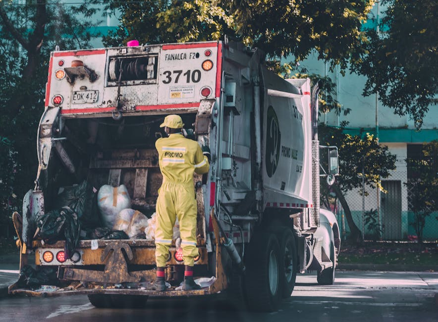 A worker dressed in a yellow high-visibility jumpsuit and helmet is seen near the back of a large waste collection truck, which is parked on a street with a row of trees and modern buildings in the background. The rear of the truck is open, revealing a collection of black and white plastic bags filled with various waste materials, some showing signs of recent collection activity. The truck's surface is slightly dirty, indicating it has been used for waste collection, with visible hoses and hydraulic components on the side. The worker appears to be checking or managing the waste load, demonstrating an active scene related to rubbish removal services. The environment suggests an urban area where private waste collection, such as that by Waste Clearance Merton, is undertaken as an alternative to local authority services, supporting a professional on-site clearance operation essential for household waste management.