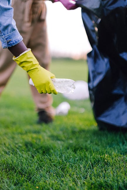 A group of volunteers wearing light blue t-shirts are participating in a litter pick-up activity in a wooded area during daylight. They are moving along a dirt path, each holding large blue trash bags filled with collected rubbish, including plastic bottles and cans. One person near the foreground is bending down to pick up litter, while others further back are actively collecting debris and placing it into their bags. The scene is set amidst tall trees with leaves in shades of green, indicating a natural outdoor environment. Soft sunlight filters through the tree canopy, creating a calm yet purposeful atmosphere. The volunteers are dressed casually with gloves for protection, and some are wearing sneakers for comfort. This image exemplifies community-led efforts to remove waste from natural areas, supporting sustainable waste management outside of formal municipal collections, with a focus on environmentally responsible rubbish removal and local cleanup initiatives.