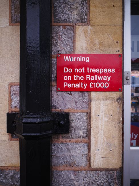 A red warning sign attached to a brick wall next to a metal pipe indicates a cautionary message. The sign reads 'Warning Do not trespass on the Railway Penalty £1000' in white text, emphasizing the prohibition against unauthorized access near railway property. The wall features a mix of textured stone and smooth yellowish bricks, with visible mortar lines. To the left, a black metal pipe runs vertically along the wall, secured by brackets, with a rough, glossy finish. In the background, part of a building with a glass window reveals what appears to be an indoor retail space, adding context to an urban environment. The lighting is natural, highlighting the textures of the bricks, mortar, and metal surface. This scene pertains to private property or infrastructure security, and waste clearance services like Waste Clearance Merton may encounter such signage when performing rubbish removal or site clearance near railway sites or restricted areas, emphasizing the importance of respecting local restrictions for safe and lawful disposal.