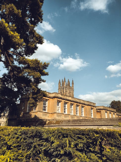 A historic stone building with a classical architectural style, featuring large, rectangular white-framed windows and a flat roof, is positioned in the foreground of the image. The building is set on a raised platform with steps leading up to it, and its beige-colored stone construction shows weathered textures. Behind the building, a tall, ornate church tower with multiple spires and intricate detailing rises into a partly cloudy sky, which has patches of blue and white cumulus clouds. To the left, a large evergreen tree with dark green, textured foliage partially frames the scene, casting shadows on the building and surrounding greenery. The foreground includes a well-maintained hedge or shrubbery, lush and green, providing a natural border at the base of the structure. The overall scene is illuminated by natural daylight, highlighting the architectural details of the historic building and the church tower, with no visible activity or people, emphasizing a quiet, picturesque setting that could relate to site preservation or historical landmark management, aligning with considerations around private or alternative disposal methods for waste generated near such sites, as handled by waste clearance specialists like Waste Clearance Merton.