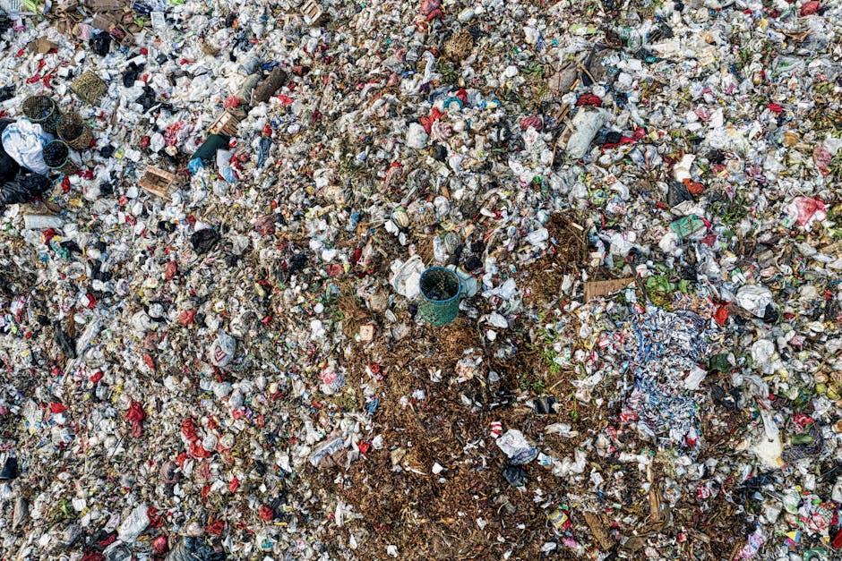 An extensive aerial view of a large landfill site showing a densely packed expanse of mixed waste materials. The heap contains a variety of discarded items such as crumpled plastic bags, paper waste, food containers, cardboard boxes, and textiles, all in different colors including white, red, blue, and green. There are sections of compacted rubbish interspersed with patches of exposed soil and dirt, with no clear organization or binarity. A teal-colored plastic bucket is partially buried within the waste, suggesting some intervention or effort to manage the refuse at this site. The surrounding environment appears to be an open area, with no visible fencing or containment features, indicative of a typical unmanaged waste dump. This scene highlights the importance of private waste handling options in areas where independent rubbish collection is necessary, showcasing the scale of unregulated waste accumulation that companies like Waste Clearance Merton can address through their rubbish removal services.