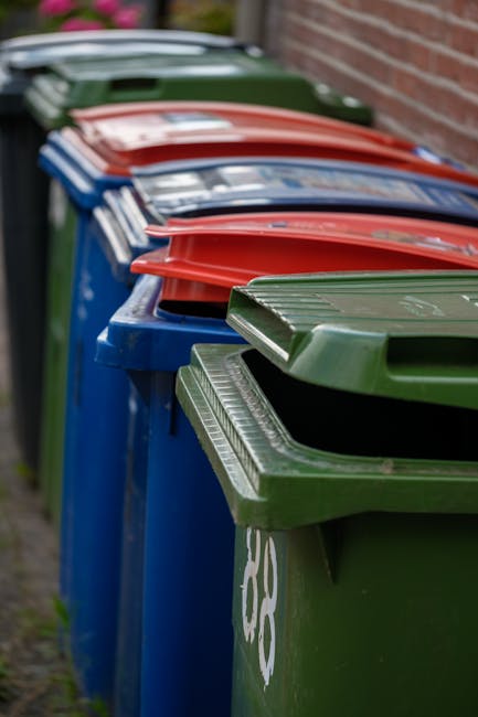 A row of seven wheelie bins lined up along a pavement next to a brick wall. The bins are made of durable plastic with textured, ribbed lids, and are in various colours including green, red, blue, and dark green. The green bin at the front has a white recycling symbol on its side, indicating it is designated for recyclable waste, while the others are plain. The bins are positioned close together, extending into the background, and are used for private rubbish disposal, potentially arranged for collection or removal by waste management services such as Waste Clearance Merton. The scene is set outdoors in natural lighting, with a slightly blurred background showing elements of a residential area, suggesting this is a typical site for waste storage prior to collection, emphasizing the importance of organized refuse management in accordance with local or private waste handling services.