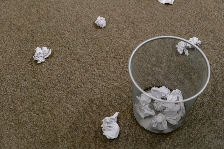 A clear cylindrical waste bin made of transparent plastic stands on a brown carpeted floor, filled with crumpled white paper. Several additional crumpled sheets of white paper are scattered across the floor around the bin, some positioned close to its base and others farther away. The paper appears to be standard letter-sized sheets that have been heavily crumpled, exhibiting irregular, wrinkled textures. The environment suggests an indoor setting, likely an office or indoor workspace with minimal background elements visible. The scene emphasizes a typical small-scale waste collection point for paper waste, reflecting an informal or private disposal context rather than a commercial or industrial rubbish removal. The lighting in the scene is neutral and even, with no prominent shadows or highlights, providing a straightforward, neutral view of the waste disposal area, which aligns with independent rubbish handling services like those offered by Waste Clearance Merton.
