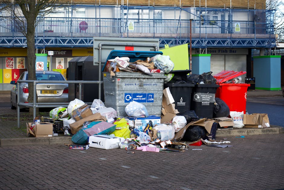 A large collection of overflowing rubbish bins, including a grey mixed waste container filled with discarded paper, cardboard boxes, and various packaging materials, some of which are spilling onto the surrounding paved area. To the left, there are several black and black-and-white bin bags piled on top of and beside the bins, containing household waste. A red recycling bin stands to the right of the grey container, partially obscured by the overflowing rubbish. Additional cardboard boxes and plastic bags are scattered across the pavement, some flattened and others intact, along with miscellaneous small items like paper and packaging debris. In the background, a building with a blue metal scaffolding structure, covered in warning signs, is visible, along with a row of shopfronts with signage indicating different retail outlets. A parked car is positioned behind a metal railing in front of the bins, and a leafless tree stands adjacent to the rubbish pile, suggesting a street scene in an urban or suburban area. The scene appears to be a typical location for private waste collection or rubbish clearance services, with Waste Clearance Merton potentially involved in handling such waste removals for this site.