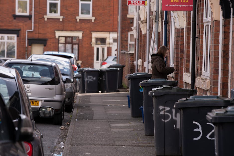 A large collection of overflowing rubbish bins, including a grey mixed waste container filled with discarded paper, cardboard boxes, and various packaging materials, some of which are spilling onto the surrounding paved area. To the left, there are several black and black-and-white bin bags piled on top of and beside the bins, containing household waste. A red recycling bin stands to the right of the grey container, partially obscured by the overflowing rubbish. Additional cardboard boxes and plastic bags are scattered across the pavement, some flattened and others intact, along with miscellaneous small items like paper and packaging debris. In the background, a building with a blue metal scaffolding structure, covered in warning signs, is visible, along with a row of shopfronts with signage indicating different retail outlets. A parked car is positioned behind a metal railing in front of the bins, and a leafless tree stands adjacent to the rubbish pile, suggesting a street scene in an urban or suburban area. The scene appears to be a typical location for private waste collection or rubbish clearance services, with Waste Clearance Merton potentially involved in handling such waste removals for this site.